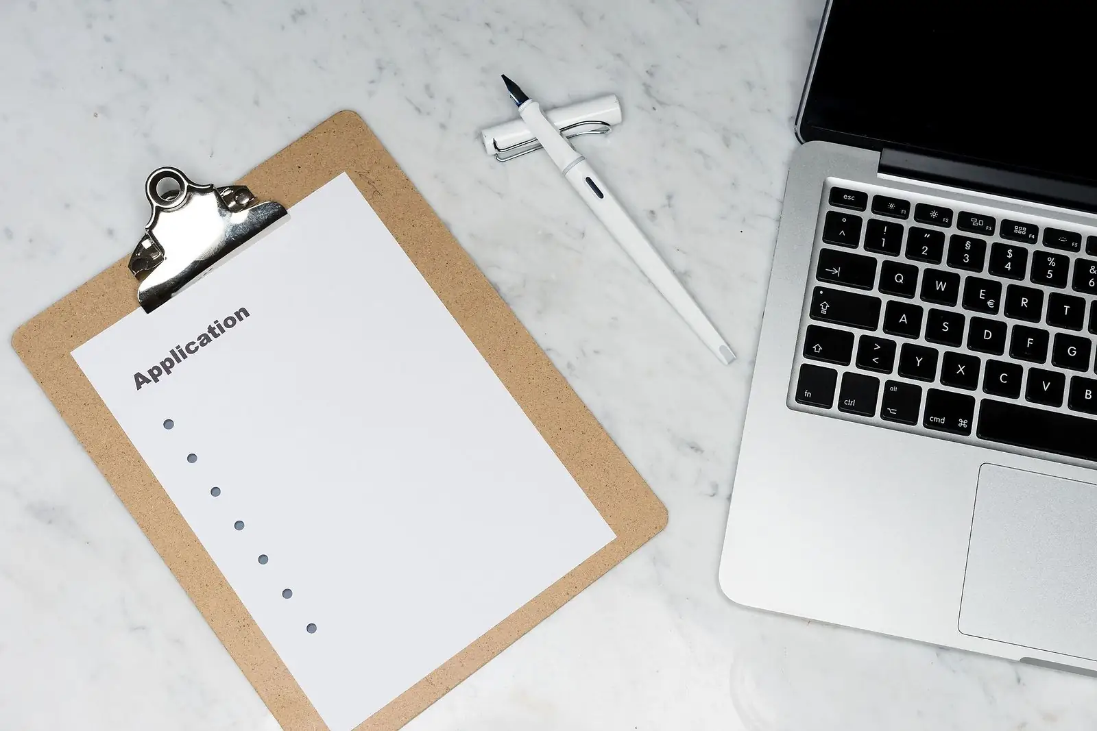 Blank application form on a clipboard beside a white pen and laptop on marble