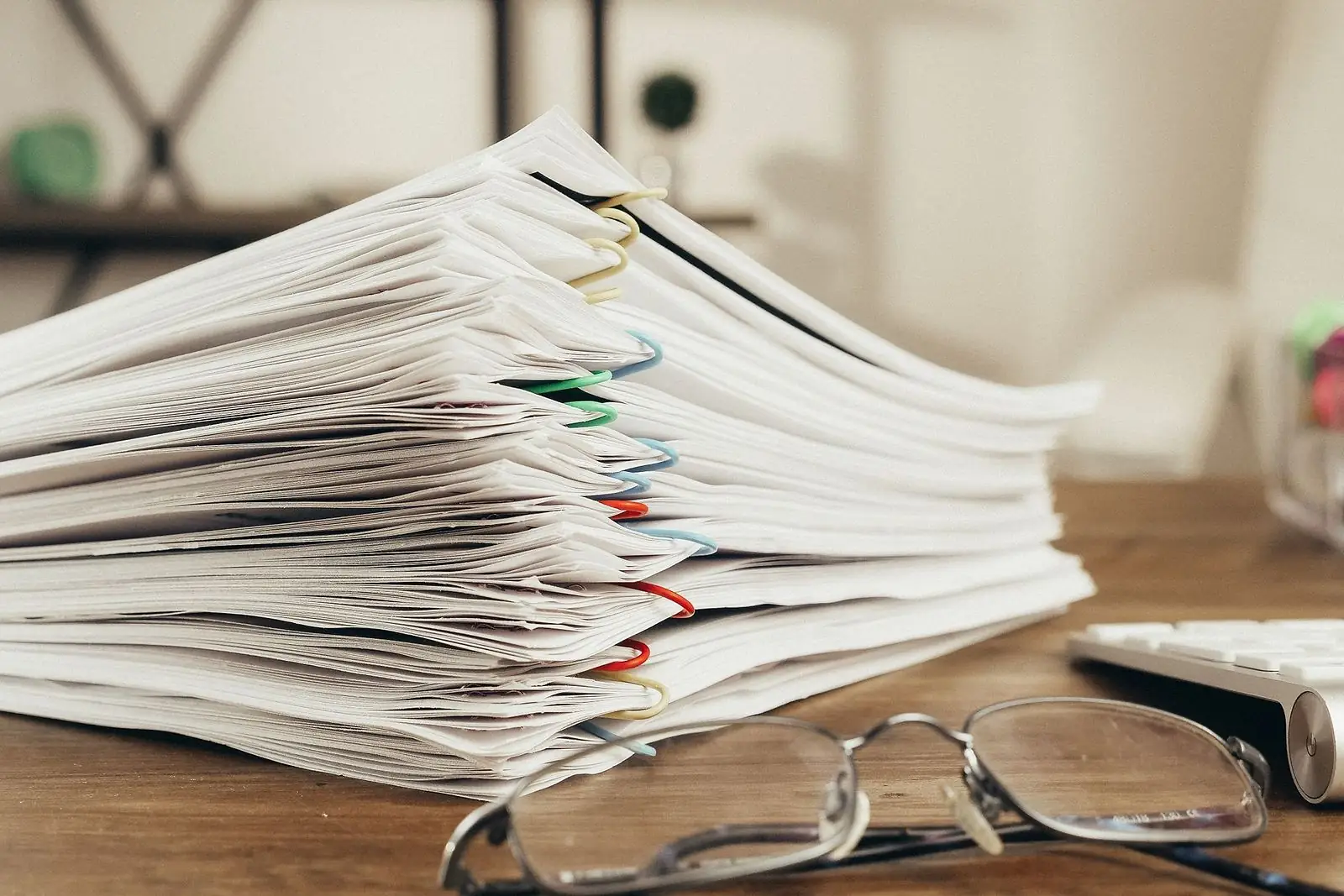 Tall stack of paper-clipped documents and reading glasses on a wooden desk