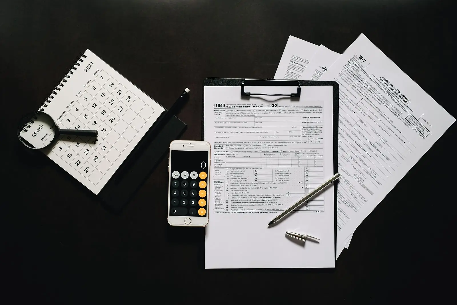 Blank tax forms on a clipboard with a calendar, magnifier and phone calculator on a dark desk