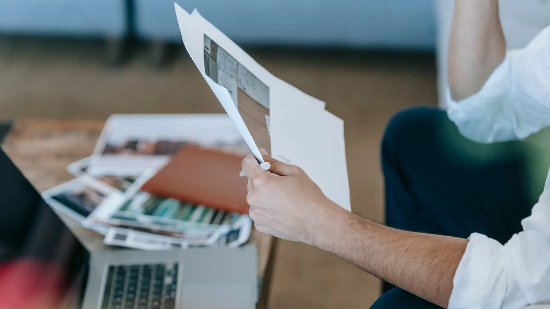 Man in a white linen shirt holding printed pages above a coffee table spread with photo prints, an open laptop in the foreground
