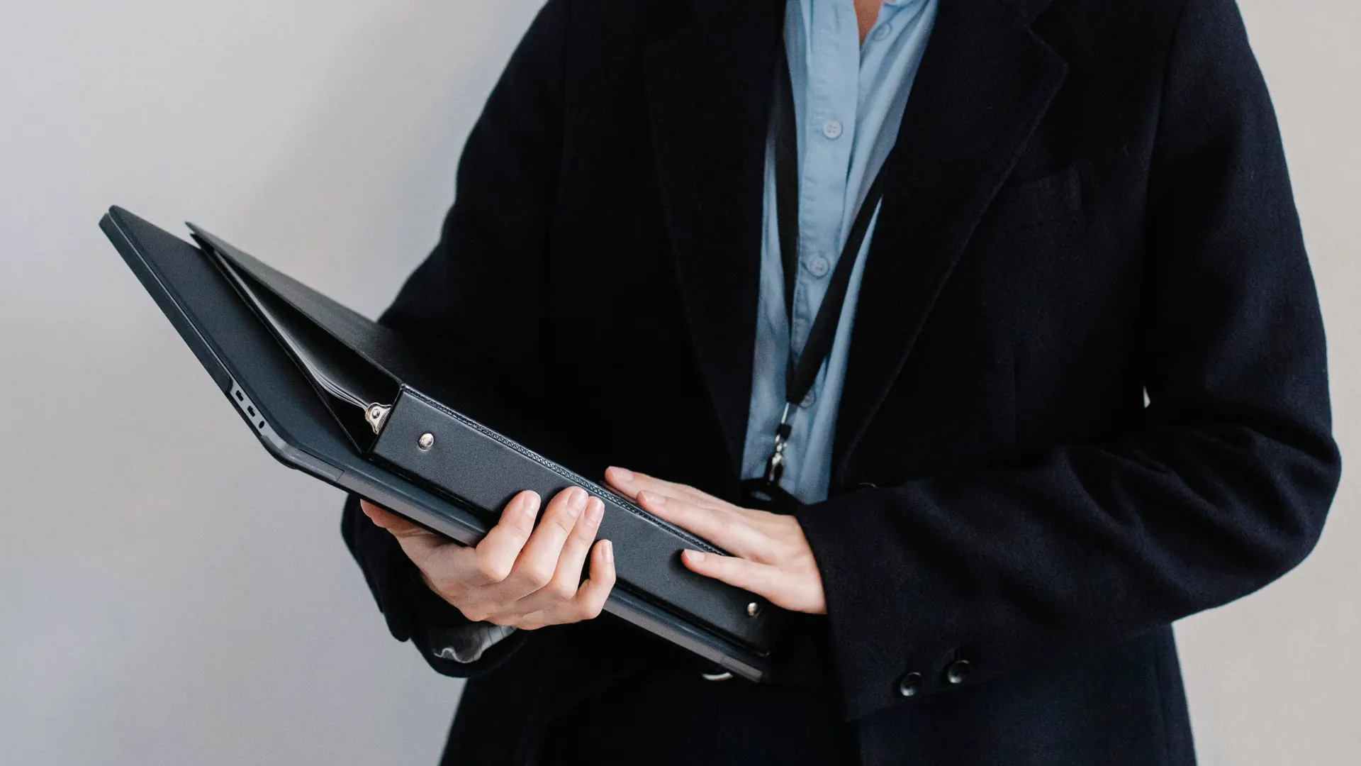 An office worker in a dark coat with an ID lanyard holds an open ring binder of records, face out of frame