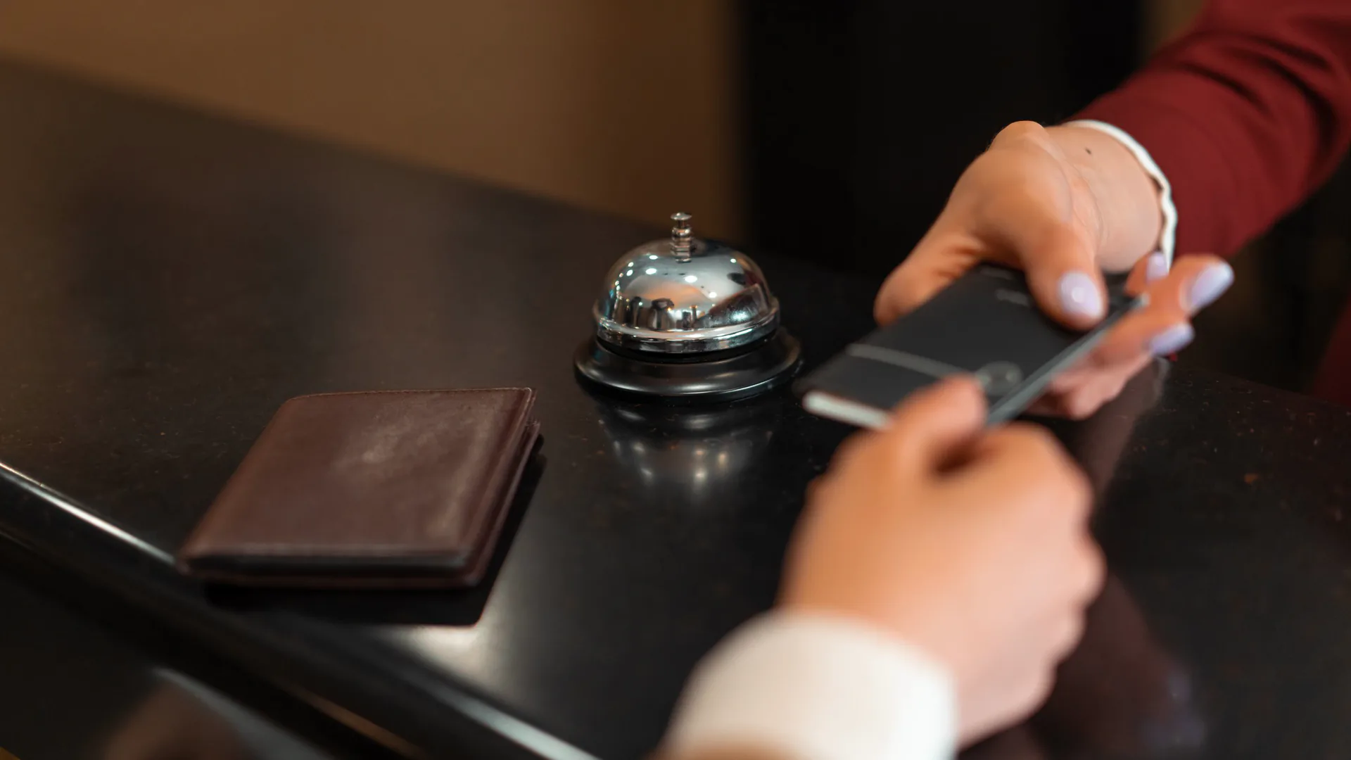 A hotel receptionist in a burgundy jacket hands a key-card folder to a guest across a dark front desk beside a service bell