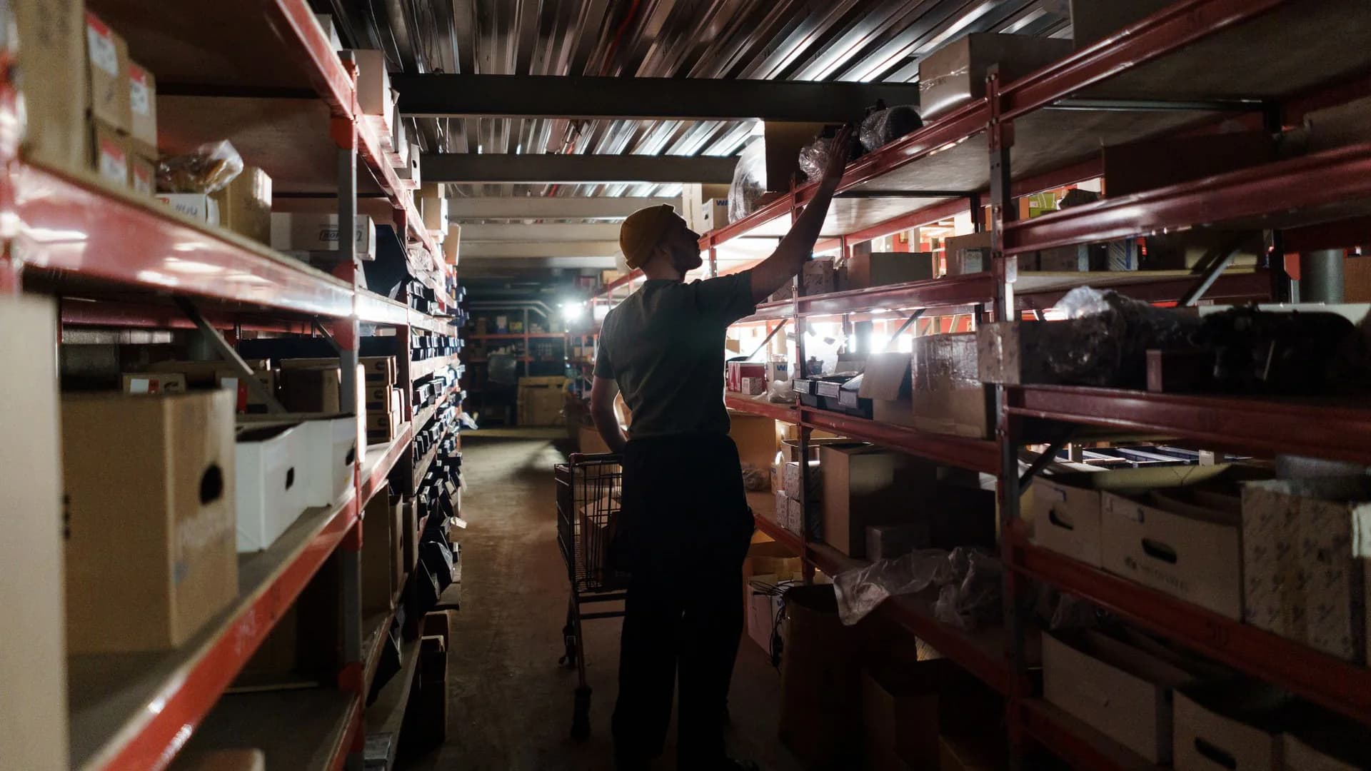 A warehouse worker seen from behind reaches up to a high shelf in a narrow aisle of red metal racks stacked with boxes, a trolley at his side