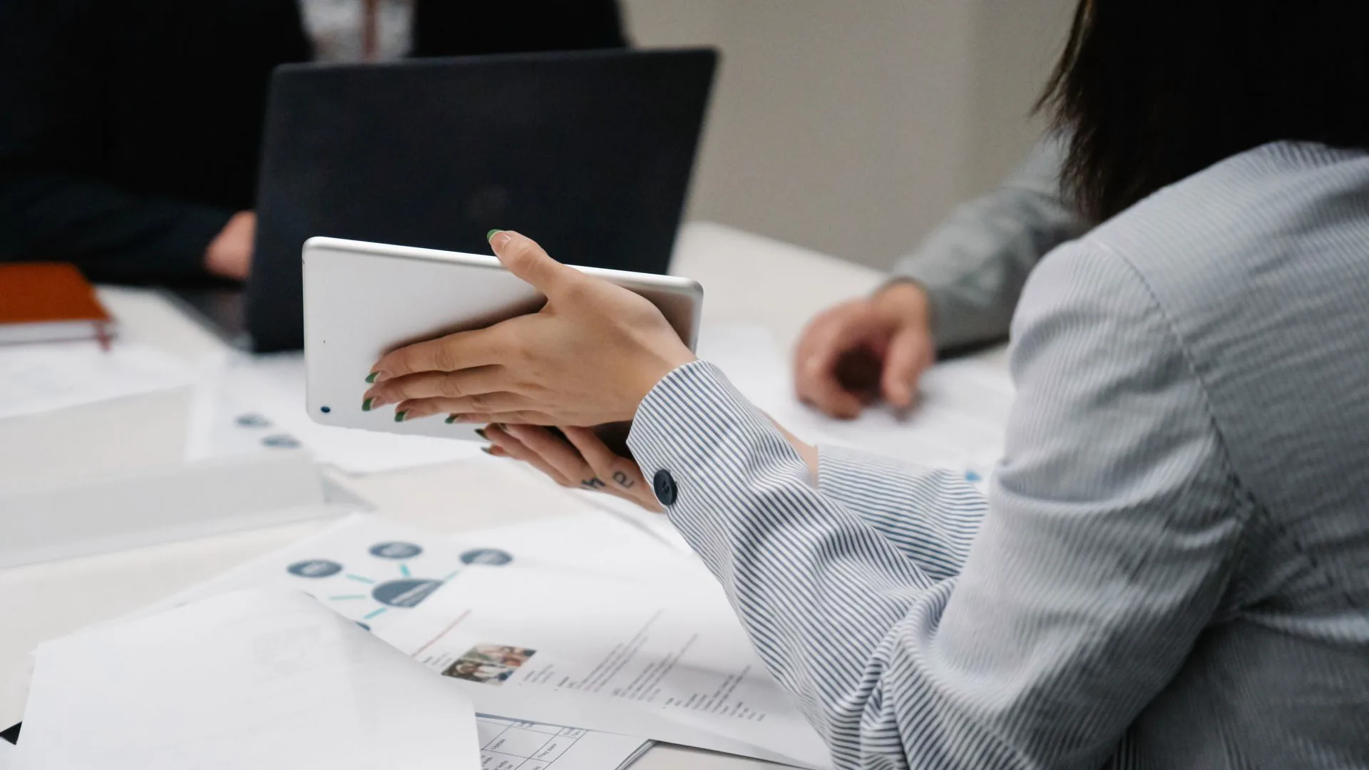 Woman seen from behind holding up a tablet across a meeting table covered with printed charts, a laptop and colleagues' hands