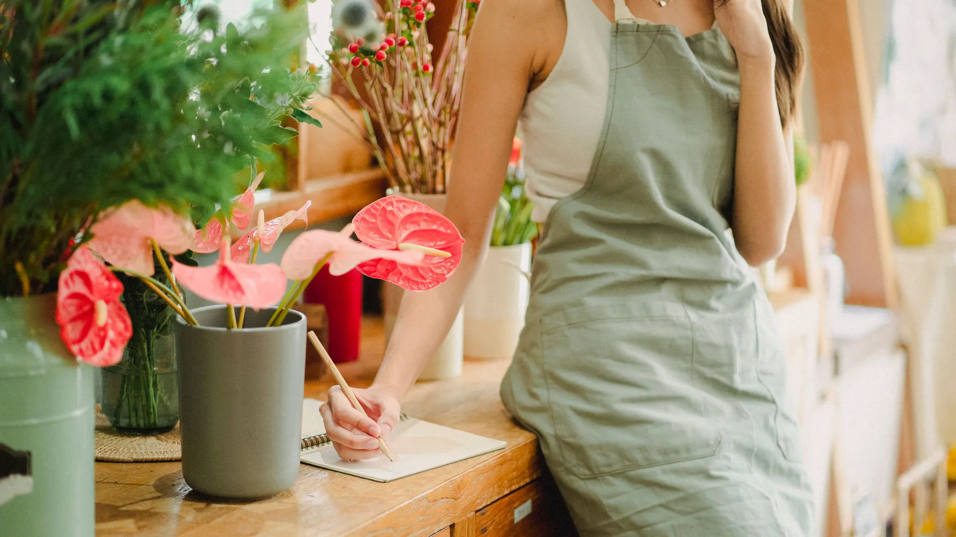 Florist in a sage apron leaning on a wooden shop counter, writing in a notebook beside pots of pink anthurium flowers